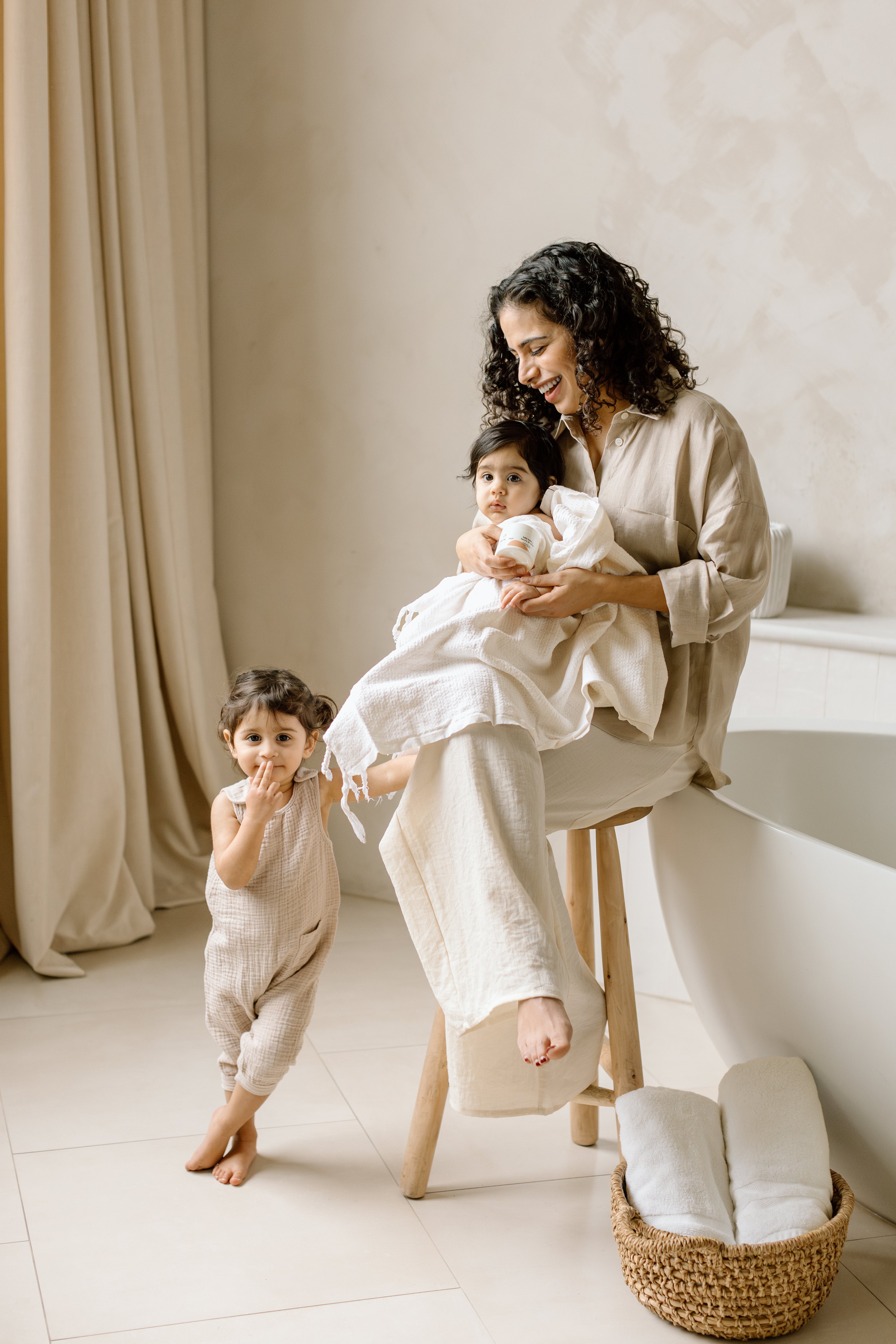 Toddler girl standing next to parent seated holding baby wrapped in towel in bathroom