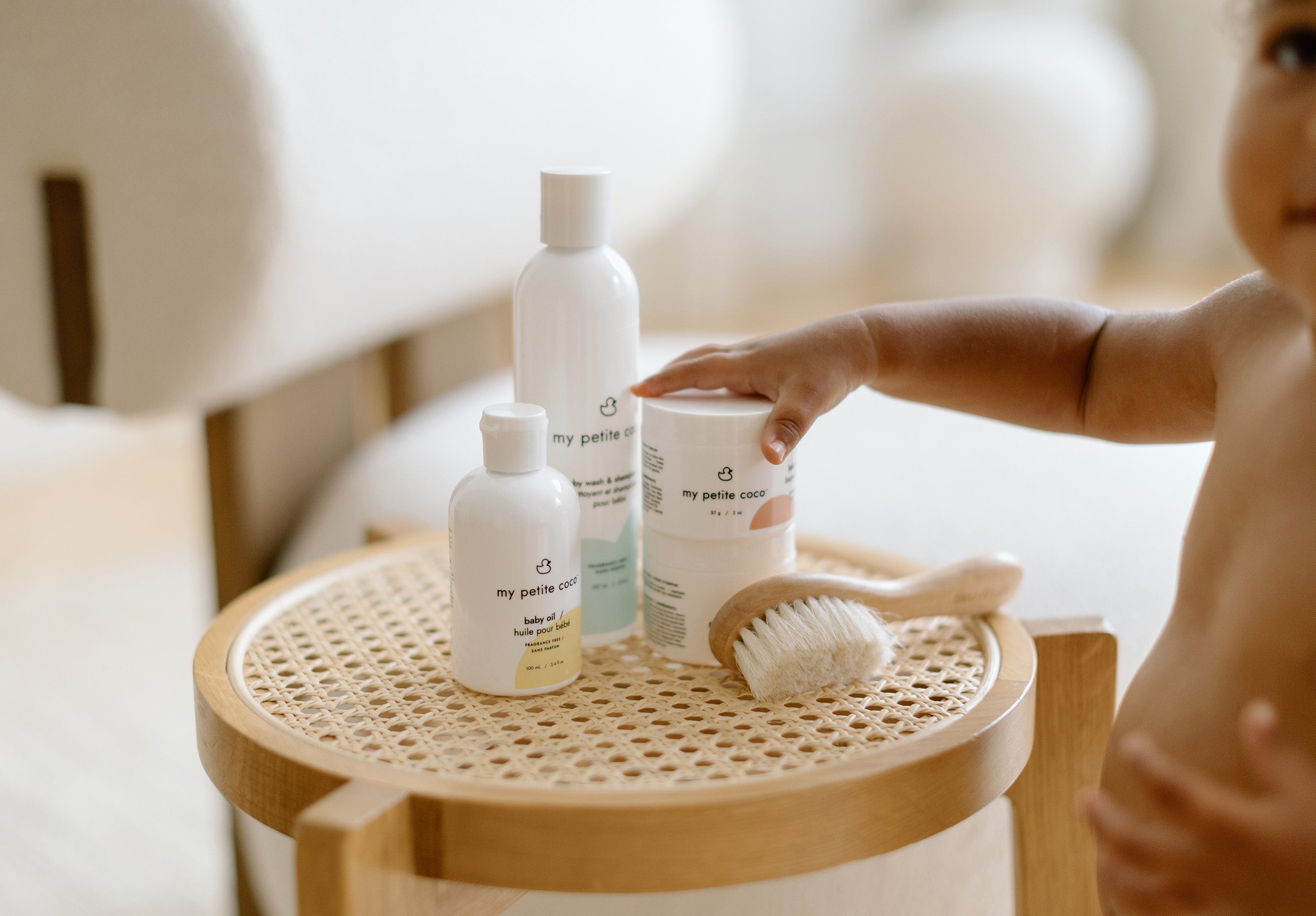Baby reaching for fragrance-free baby balm on a wicker side table with baby skincare products
